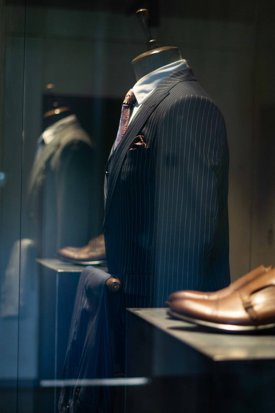 Detaljebillede fra en SUIT CLUB-butik af en mannequin set fra siden, iført navy nålestribet blazer, hvid skjorte og slips. Brune sko ses i både forgrunden og baggrunden.