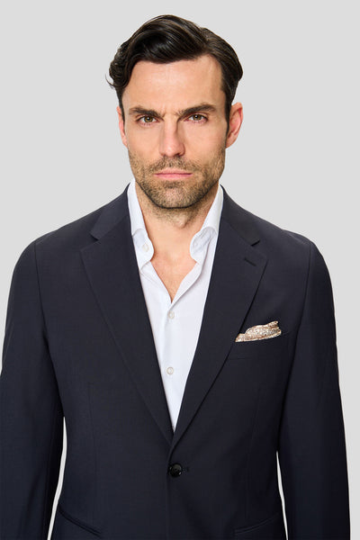 Mandlig model iført navy Premium-blazer stylet med hvid skjorte og lommeklud står foran en grå baggrund og ser mod kameraet.