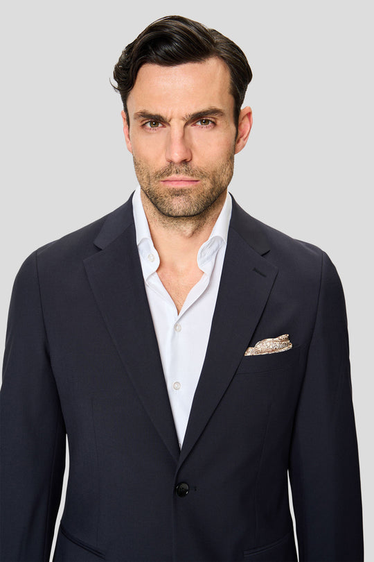 Mandlig model iført navy Premium-blazer stylet med hvid skjorte og lommeklud står foran en grå baggrund og ser mod kameraet.