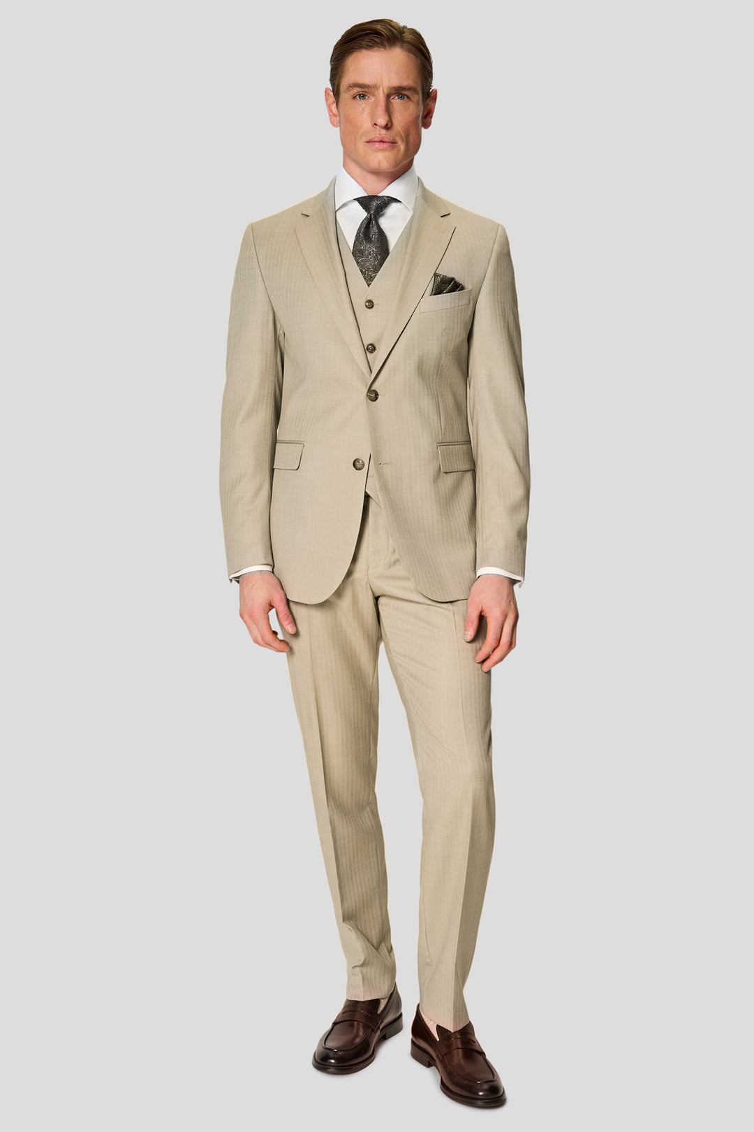 Mandlig model iført beige Heritage three-piece jakkesæt med sildebensmønster stylet med hvid skjorte, grønt slips, matchende lommeklud og sorte loafers står foran en grå baggrund og ser mod kameraet.